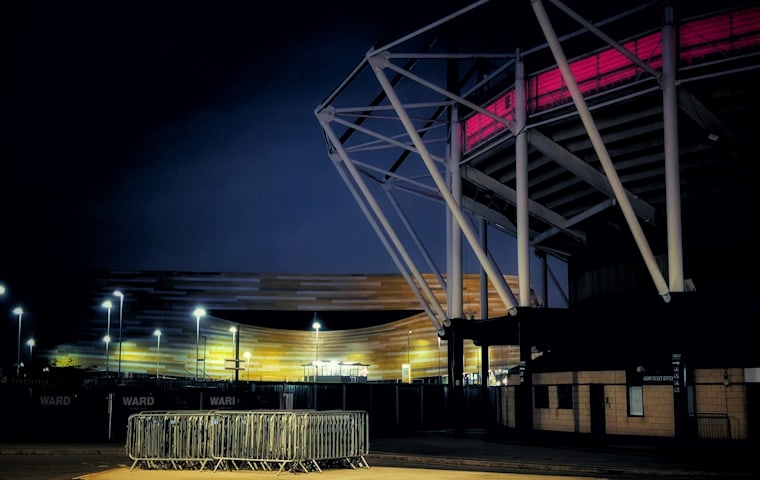 A floodlit stadium at night with crowd control barriers stacked outside